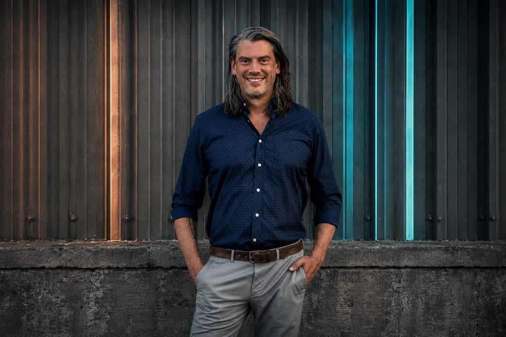 Zach Arrington, standing in front of a corrugated metal wall lit in cyan and amber.